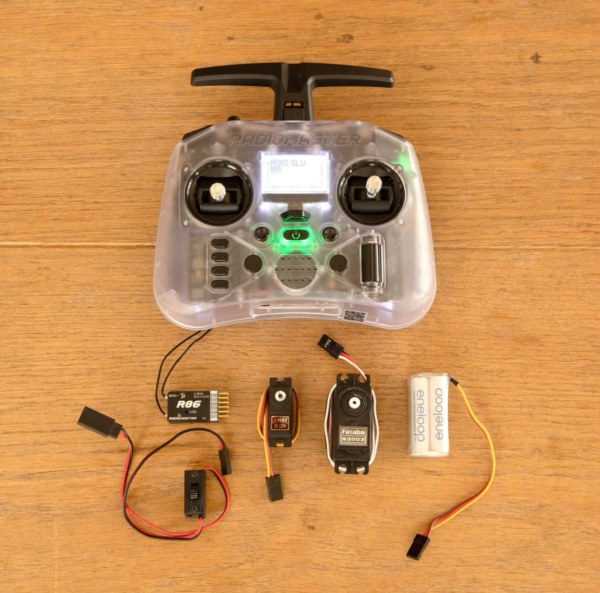 Equipo de control remoto para un yate modelo dispuesto sobre un suelo de madera. La configuración incluye un transmisor Pocket de RadioMaster transparente con su pantalla mostrando “RG65 SLU,” un receptor R86 con cables de antena, un servo Futaba S3003, u