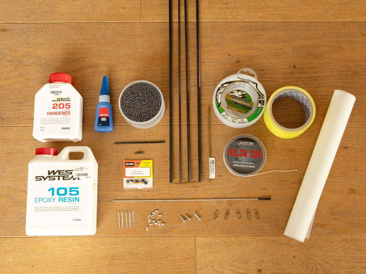 Una selección de materiales para la construcción de yates de modelo dispuestos ordenadamente sobre un suelo de madera. Los artículos incluyen: resina epóxica West System 105 (1L) y endurecedor rápido 205 (200ml), una botella de pegamento super Loctite, un