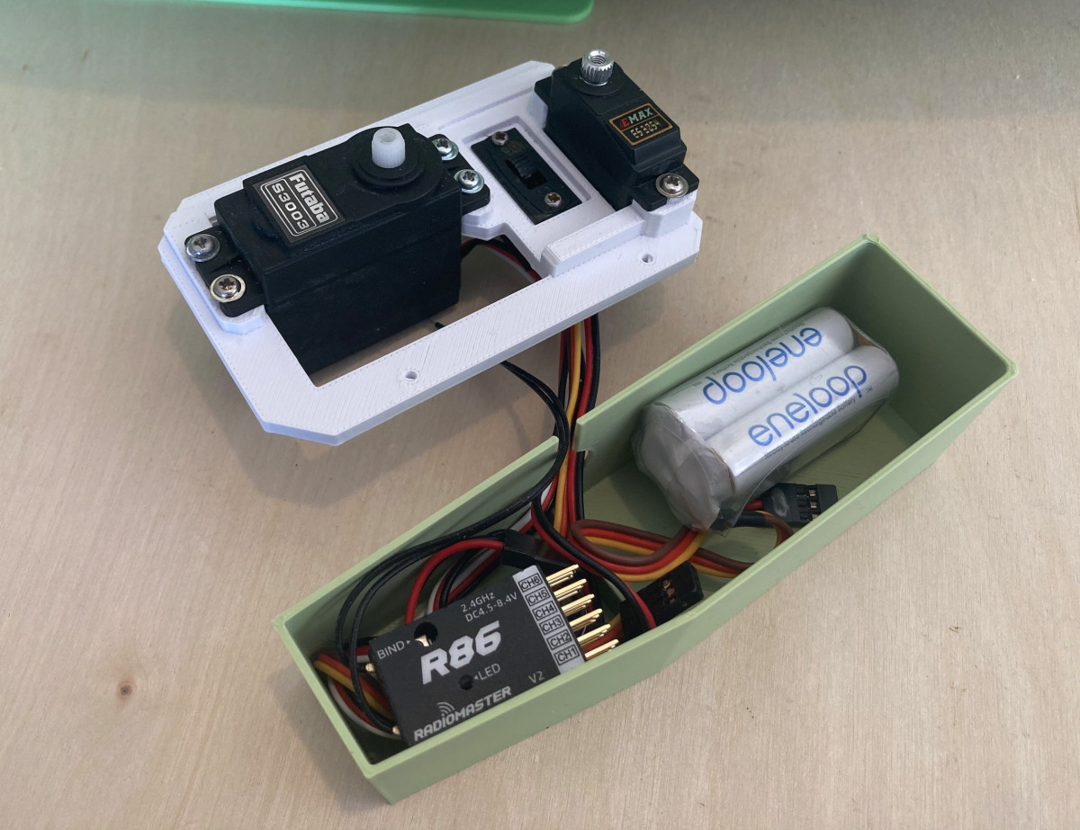 Acercamiento de una instalación de control remoto de modelo de yate. Una bandeja de servo impresa en 3D de color blanco sostiene un servo Futaba S3003 y un servo EMAX ES08MA II con un interruptor de encendido/apagado entre ellos. Junto a ello, una bandeja