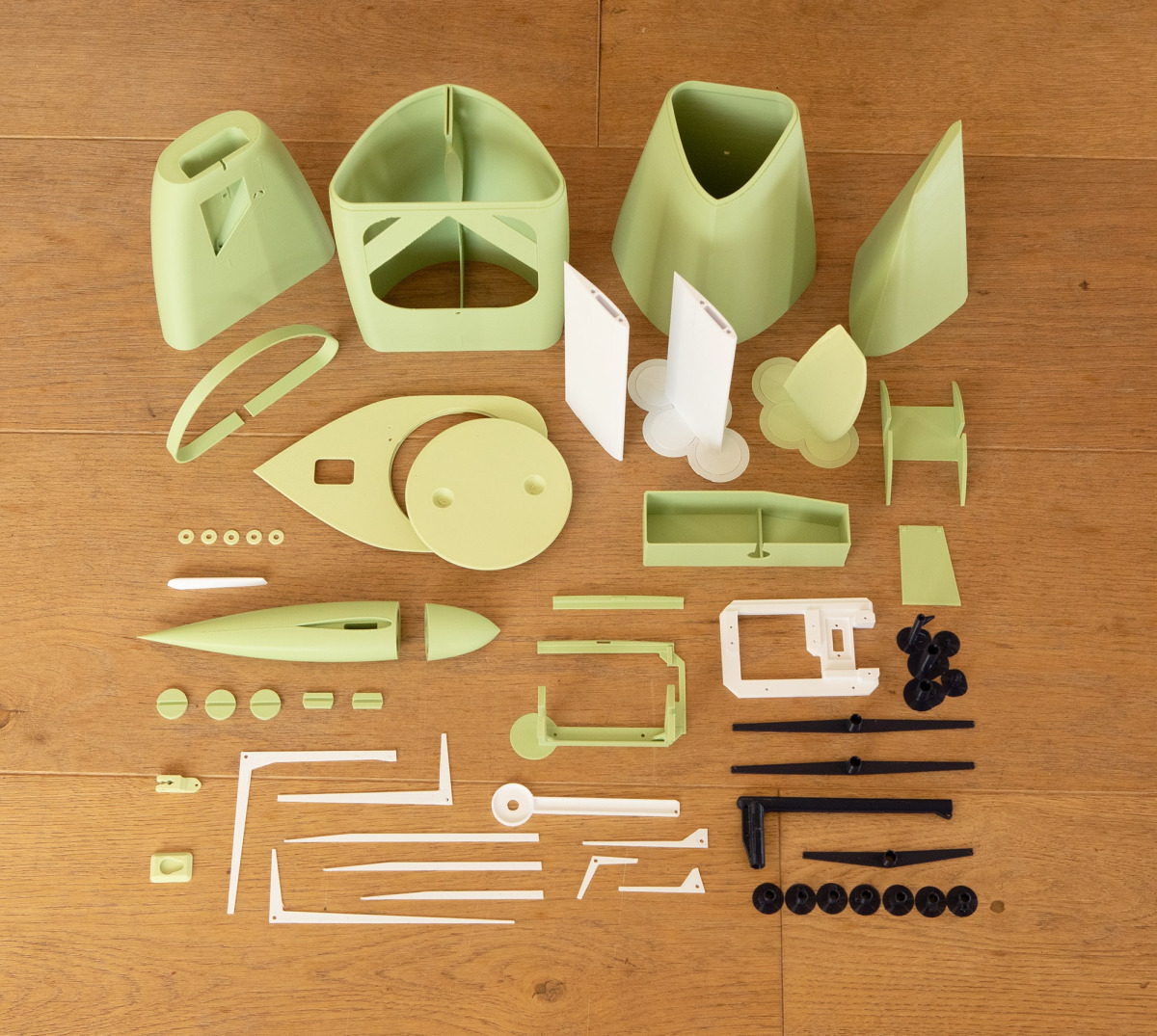 Un conjunto completo de piezas de modelo de yate impresas en 3D en plástico verde claro, blanco y negro, dispuestas sobre un suelo de madera. El conjunto incluye secciones del casco, aletas de quilla y mitades de bulbo, accesorios de cubierta, bandeja de 