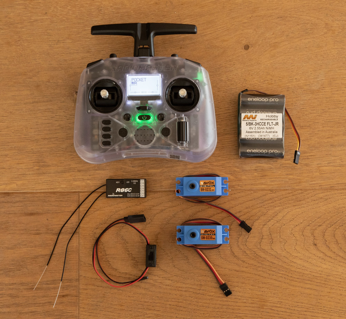 Ein Satz von Funksteuerungskomponenten, die auf einem Holzboden angeordnet sind. Die Gegenstände umfassen: einen transparenten RadioMaster Pocket RC-Sender mit einem Display, einen R86C-Empfänger mit Antennenkabeln, zwei blaue Savox SW-0231MG-Servos mit a