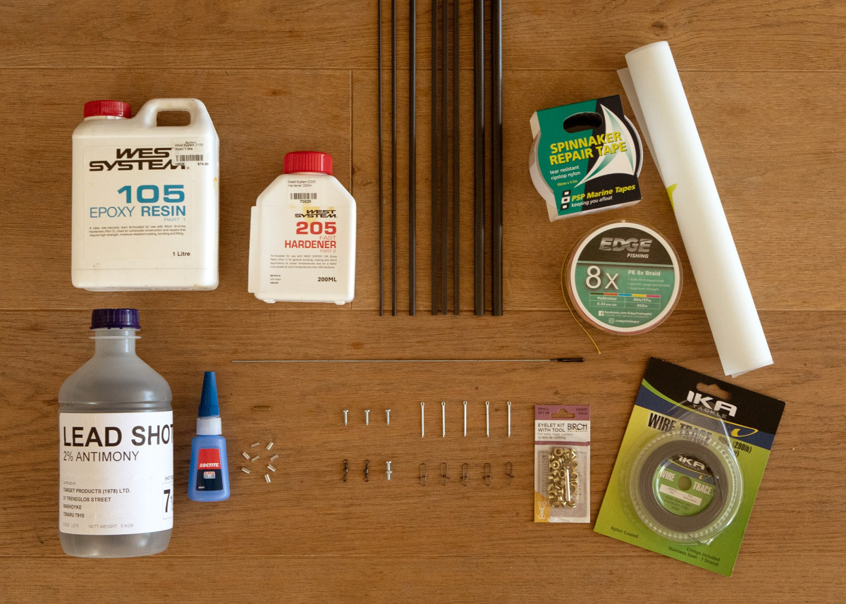 Ein ordentlich angeordnetes Set von Materialien zum Bau von Modellyachten, das auf einem Holzboden ausgelegt ist. Die Gegenstände umfassen: einen 1-Liter-Behälter mit West System 105 Epoxidharz und eine 200-ml-Flasche mit West System 205 Schnellhärter, ei