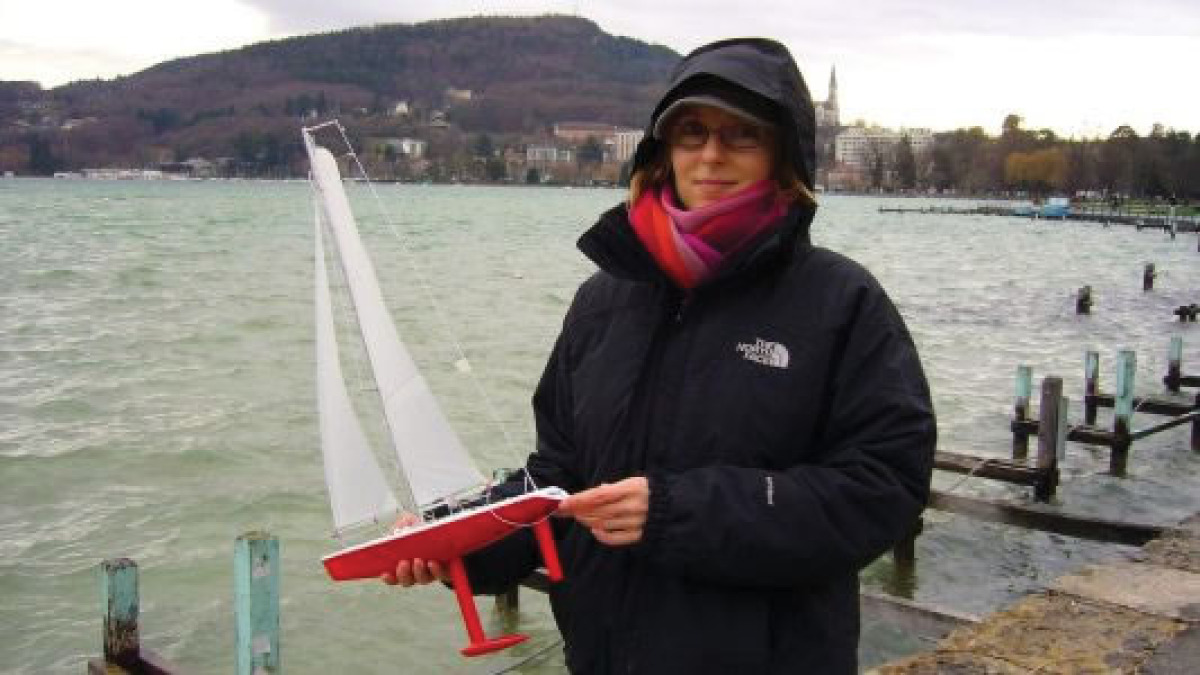 A woman holding a tiny radio controlled sailboat, a Racing Sparrow 375mm. It looks cold as she is wearing a thick jacket with a hood and scarf.