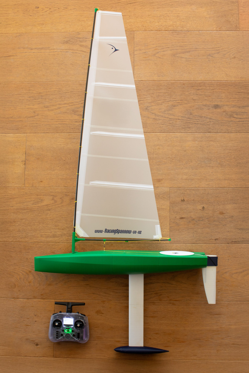 RS Ranger 695 3D printed RC sailboat in green with white sail, shown fully assembled with keel and rudder, standing upright on wooden floor next to a radio controller.