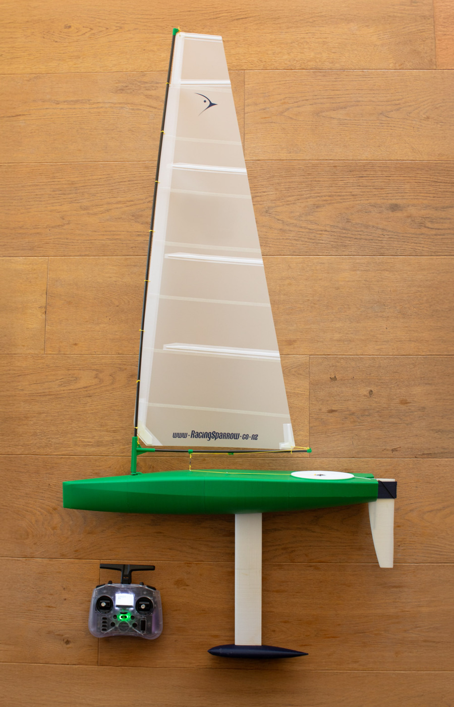 RS Ranger 695 3D printed RC sailboat in green with white sail, shown fully assembled with keel and rudder, standing upright on wooden floor next to a radio controller.